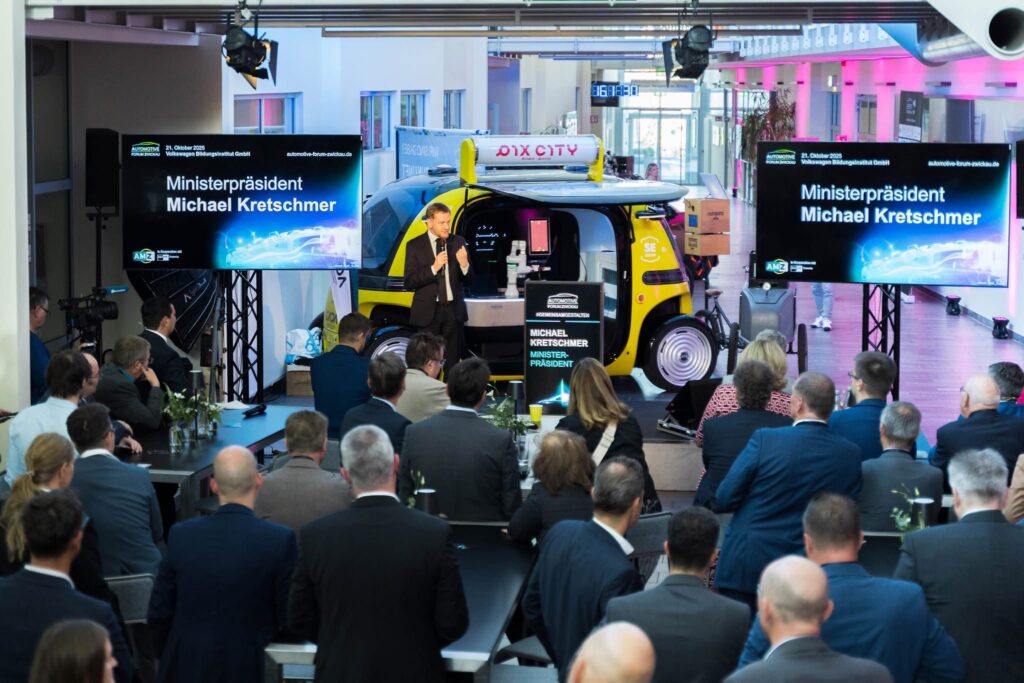 Minister President Michael Kretschmer speaks at the Automotive Forum Zwickau 2025 in front of a yellow autonomous vehicle by staff-eye, while the audience listens attentively. Large screens display his name and the venue, Volkswagen Bildungsinstitut GmbH.<br>