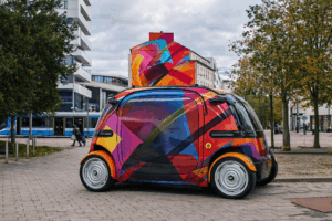 Farbenfrohes autonomes Fahrzeug mit MadC-Kunstdesign steht auf einem Stadtplatz, im Hintergrund ein großes urbanes Wandgemälde und eine Straßenbahn.